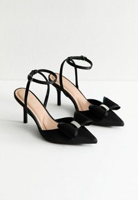 Talons aiguilles à bout pointu noirs avec des brides de cheville et des nœuds décoratifs ornés de strass sur une surface blanche.