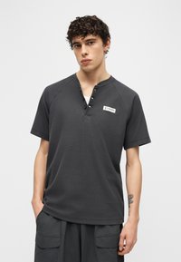 Νεαρός άνδρας με σγουρά μαλλιά, φορώντας γκρι σκούρο κοντομάνικο πουκάμισο henley με λογότυπο Columbia και αντίστοιχο παντελόνι τύπου cargo.