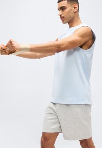 Άνδρας με ανοιχτό μπλε μπλουζάκι χωρίς μανίκια και γκρι σορτς τεντώνει τον καρπό του με ελαστική ταινία μπροστά σε απλό λευκό φόντο.