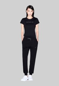 Zwarte katoenen t-shirt met verhoogd logo, gecombineerd met zwarte joggers met een trekkoord in de taille en logodetails, aangevuld met witte sneakers.
