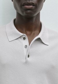Polo shirt grigio chiaro con una maglia testurizzata, due bottoni sul posto dei bottoni e un design del collo classico. In primo piano su colletto e scollatura.