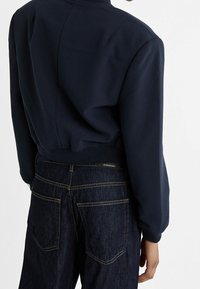 Navy bomber jacket with a ribbed collar and hem, featuring a smooth texture. Paired with dark denim jeans showcasing patch pockets.