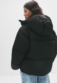 Crna puffer jakna s kapuljačom, koja ima kvadrirani dizajn i uočljivu debljinu, uparena s svijetloplavim trapericama.