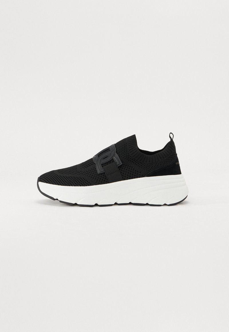 Baskets noires de sport avec une tige en tricot, dotées d'une sangle bien visible. Semelle épaisse blanche avec une finition texturée et des accents noirs.