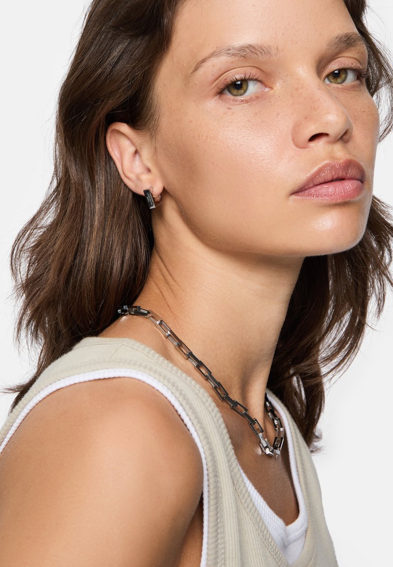 Schwarze Metalohrringe und eine Kettenhalskette werden mit einem hellbeigen ärmellosen Oberteil getragen. Die Halskette besteht aus miteinander verbundenen rechteckigen Formen.