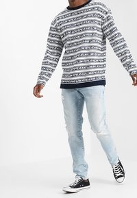 Pitkähihaista tummansinistä ja valkoista paitaa toistuvalla tekstikuviolla, yhdistettynä vaaleansinisiin kuluneisiin farkkuihin ja mustiin lenkkareihin.