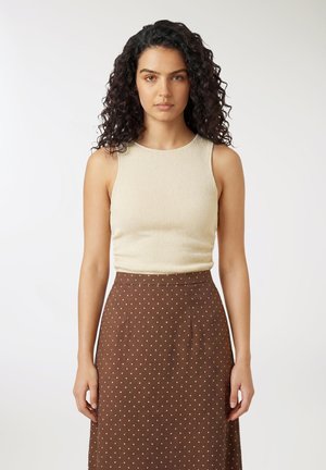 Donna con capelli ricci scuri che indossa una canottiera beige e una gonna marrone a vita alta con piccoli pois bianchi, in piedi davanti a uno sfondo bianco.