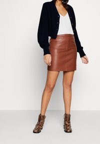 Brauner Minirock aus Leder, kombiniert mit einem weißen V-Ausschnitt-Top und einer marineblauen Strickjacke. Accessoires sind gemusterte Stiefeletten.