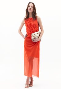 Strak aansluitende oranje maxi-jurk met geplooide details, doorzichtig onderste gedeelte en een soepele textuur. Gecombineerd met een metallic gouden clutch.
