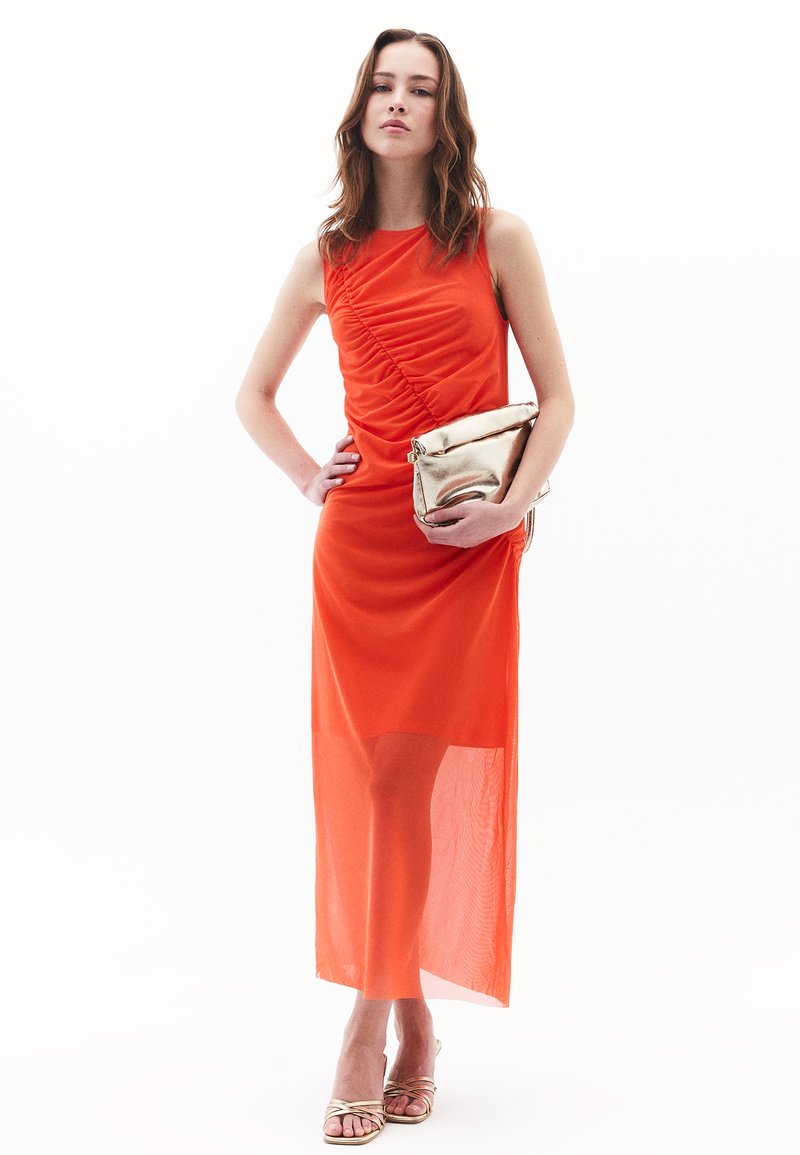 Strak aansluitende oranje maxi-jurk met geplooide details, doorzichtig onderste gedeelte en een soepele textuur. Gecombineerd met een metallic gouden clutch.