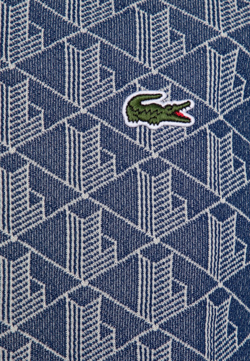 Marine stof met een geometrisch wit patroon, voorzien van een klein groen krokodillenlogo erop geborduurd. Voelbare textuur details zichtbaar.