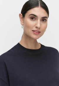 Marineblauwe gebreide trui met een ronde halslijn, geribbelde manchetten en een gladde textuur. Model heeft eenvoudige oorbellen en een neutrale make-uplook.