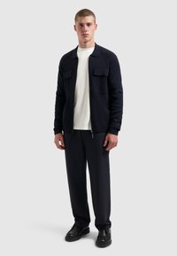 Marineblauwe jasje met kraag en twee voorzakken, gedragen over een wit shirt. Gecombineerd met donkere broek en zwarte schoenen. Glad textuur, moderne pasvorm.