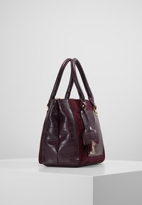 Bordeaux håndtaske med krokodillepræget kunstlæder, dobbelte håndtag og en tekstureret side. Har en aftagelig charme og guldhardware.