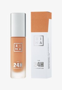 Flytande foundation i en klar glasflaska, med ett silverlock. Har en ljus persikofärg och en vit låda med koppardetaljer. Text tryckt på förpackningen.