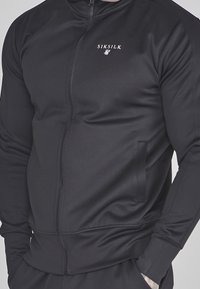 Chaqueta negra con cremallera, hecha de tela suave, que cuenta con un cuello alto, bolsillos laterales y un pequeño logo en el pecho. Corte ajustado con mangas largas.