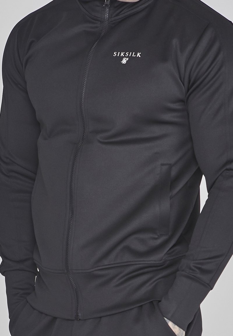 Chaqueta negra con cremallera, hecha de tela suave, que cuenta con un cuello alto, bolsillos laterales y un pequeño logo en el pecho. Corte ajustado con mangas largas.