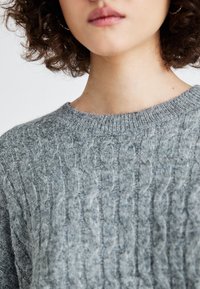 Frau mit lockigem Haar, die einen strukturierten grauen Kabelstrickpullover und Ohrringe trägt, Lippen geschlossen und Hals sichtbar.