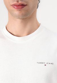 Κοντινό σε άτομο που φοράει λευκό μπλουζάκι με στρογγυλή λαιμόκοψη με το λογότυπο "Tommy Jeans" και μια μικρή κόκκινη, λευκή και μπλε σημαία στο στήθος.