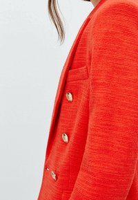 Blazer rouge vif en tissu texturé, avec des boutons dorés et une poche poitrine unique. Il présente une coupe ajustée avec un revers cranté.