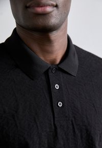 Polo nero caratterizzato da un tessuto testurizzato con un motivo geometrico, un colletto classico e tre bottoni coordinati sul colletto.