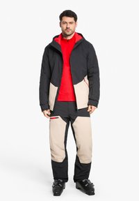 Skijacke in Schwarz und Beige, mit einem roten Innenfutter. Kombiniert mit passenden Hosen, die über Taschen und ein schlankes, wasserdichtes Design verfügen.