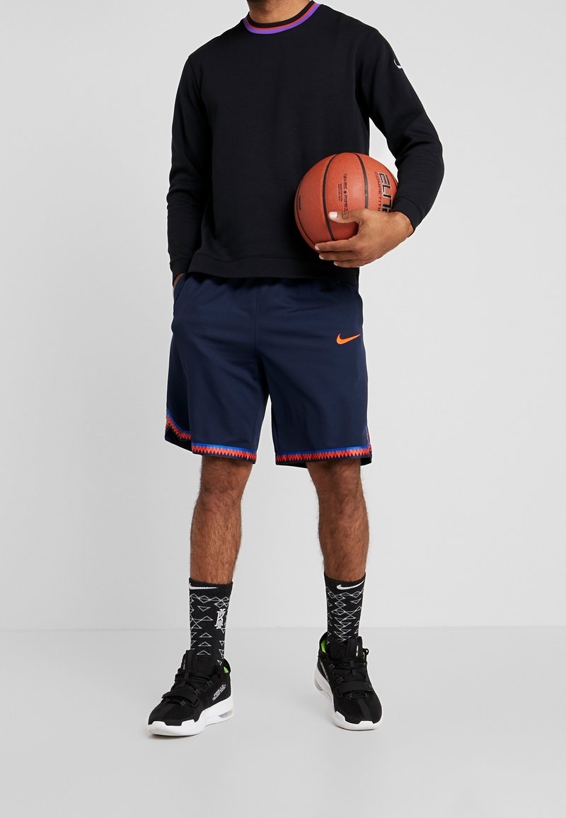 Navy Basketball-Shorts mit orangefarbenem Nike-Logo und gemustertem Saum, schwarzes Langarm-T-Shirt mit lila Kragen, strukturierte schwarze Socken, schwarze Schuhe.