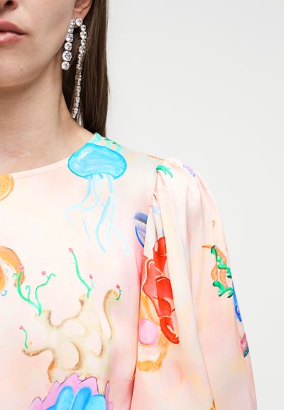 Femme portant une longue boucle d'oreille en strass et un chemisier rose pastel avec des imprimés colorés de créatures marines, dont des méduses et du corail.