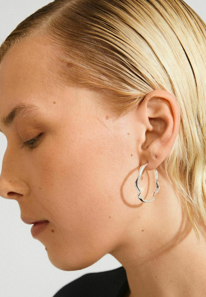 Nærbillede af en person med slicked-back blond hår, der bærer en struktureret sølvring i venstre øre.