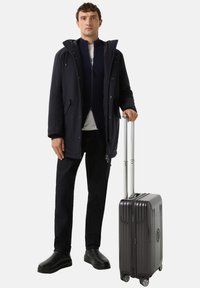 Tmavě modrá parka s kapucí, černá vrstva se zipem pod ní, černé džíny a černé boty. Drží šedý pevný kufr na kolečkách.