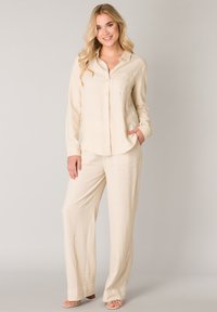 Hellbeiges Leinenhemd mit durchgehender Knopfleiste und einer einzelnen Brusttasche, kombiniert mit passenden, locker sitzenden Hosen. Glatte Textur.