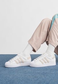 Pessoa a usar ténis Adidas Superstar brancos com riscas bege, meias brancas e calças bege claro, sentada num tapete azul.