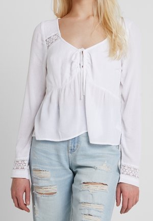 Femme portant un chemisier blanc à manches longues avec des détails en dentelle et un jean bleu clair déchiré, debout devant un fond uni.