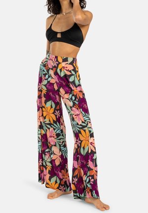 Frau trägt ein schwarzes Crop-Top und weite Hosen mit buntem Blumenmuster, steht barfuß vor einem schlichten Hintergrund.