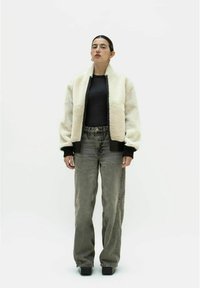 Cremefarbene Kunstpelz-Bomberjacke mit schwarzen gerippten Bündchen und Saum, kombiniert mit hoch geschnittenen grauen Jeans. Das Model steht vor einem weißen Hintergrund.