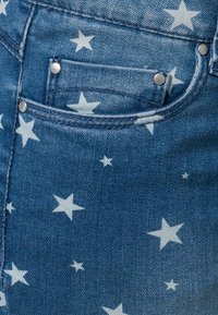 Jean en denim bleu avec un motif d'étoiles blanches, une poche avant et des rivets en métal. La texture est tissée, avec un design décontracté.