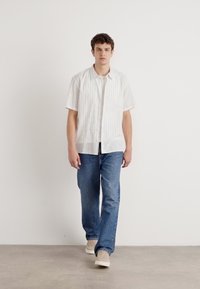 Cămașă albă cu mânecă scurtă, cu dungi verticale deschise, asortată cu blugi denim de un albastru mediu și teniși bej. Design simplu și casual.