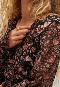 Blouse noire à motifs floraux avec des manchons froncés, présentant un motif de roses et de feuilles dans des tons de rose et de blanc. Une bague dorée sur la main ajoute une touche de détail.