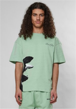 Mintgrøn bomulds T-shirt med oversized pasform, med et sort grafisk design på siden og lille logo tekst på brystet.