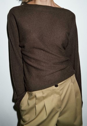 Person som bär en brun långärmad tröja inskjuten i högmidjade beige plisserade byxor, står mot en enkel bakgrund.