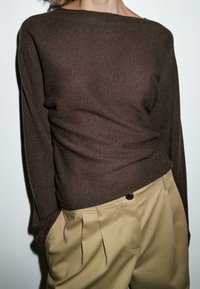 Persona che indossa un maglione lungo marrone a maniche lunghe infilato in pantaloni beige a vita alta con pieghe, in piedi contro uno sfondo neutro.
