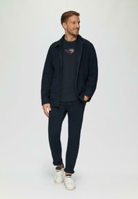 Marineblauwe jas met een knoopsluiting, gecombineerd met een bijpassend marineblauw crew neck t-shirt en broek. Witte sneakers met zwarte veters.