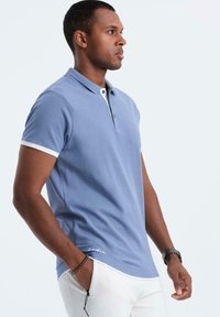 Světle modrá polo košile s krátkým rukávem a klasickým límcem, bílé detaily na rukávech a vyšívané logo poblíž spodního lemu.