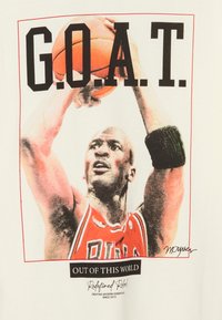 Biała koszulka z grafiką koszykarza w czerwonym stroju z napisem "G.O.A.T." oraz nadrukowanym podpisem.