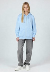 Felpa zip blu chiaro con tasca kangaroo frontale, polsini a coste e cappuccio con cordino, abbinata a jeans grigi a gamba larga e sneakers bianche.