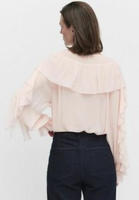 Vaaleanpunainen paita, jossa on röyhelökoristeet, pitkät hihat ja istuvat hihansuut. Paita on puettu tummien farkkujen päälle, mikä tuo esiin sujuvan ja virtaavan tekstuurin.