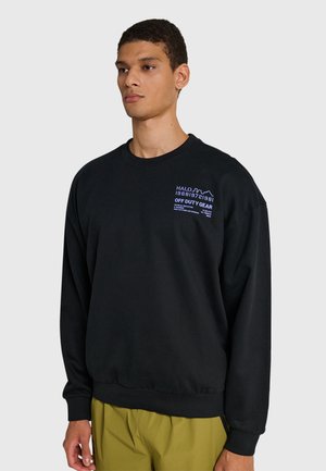 Młody mężczyzna ubrany w czarną bluzę z małym niebieskim napisem na piersi "OFF DUTY GEAR" oraz oliwkowozielone spodnie, stojący na jednolitym tle.