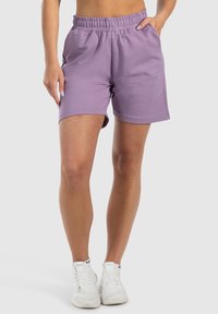 Shorts sportivi lilla con cintura elastica, tasche laterali e una texture morbida. Abbinati a scarpe sportive bianche.