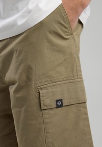 Olivi vihreät cargo-shortsit, jotka on valmistettu teksturoidusta kankaasta. Niissä on sivutasku mustalla tarralla, ommellut yksityiskohdat ja rento istuvuus.