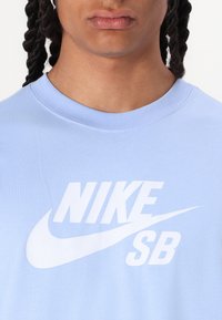 Gaiši zils kokvilnas T-krekls ar apaļu dekoltē, uz kura priekšpuses ir liels balts Nike SB logo, gluda tekstūra un īsas piedurknes.